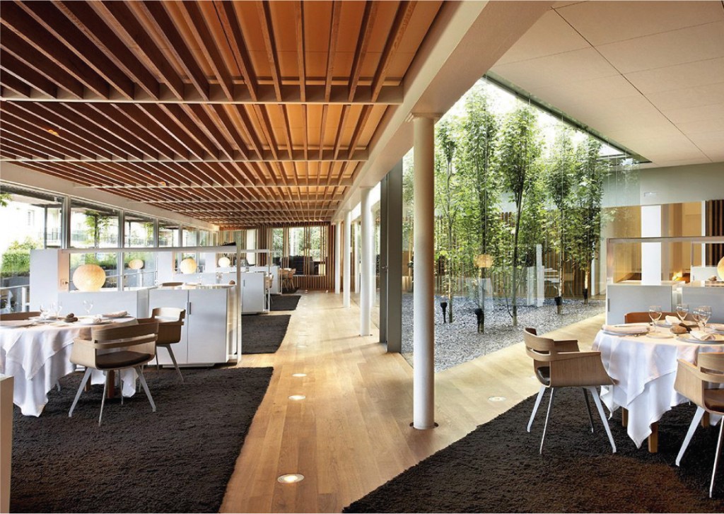 Comedor con techo de lamas de madera, mesas redondas y jardín interior tras el cristal